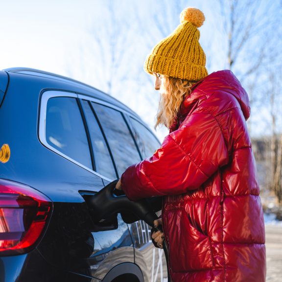Woman in red jacket and yellow hat charging her electric car at home, symbolising energy efficiency and eco-friendly electrical upgrades.